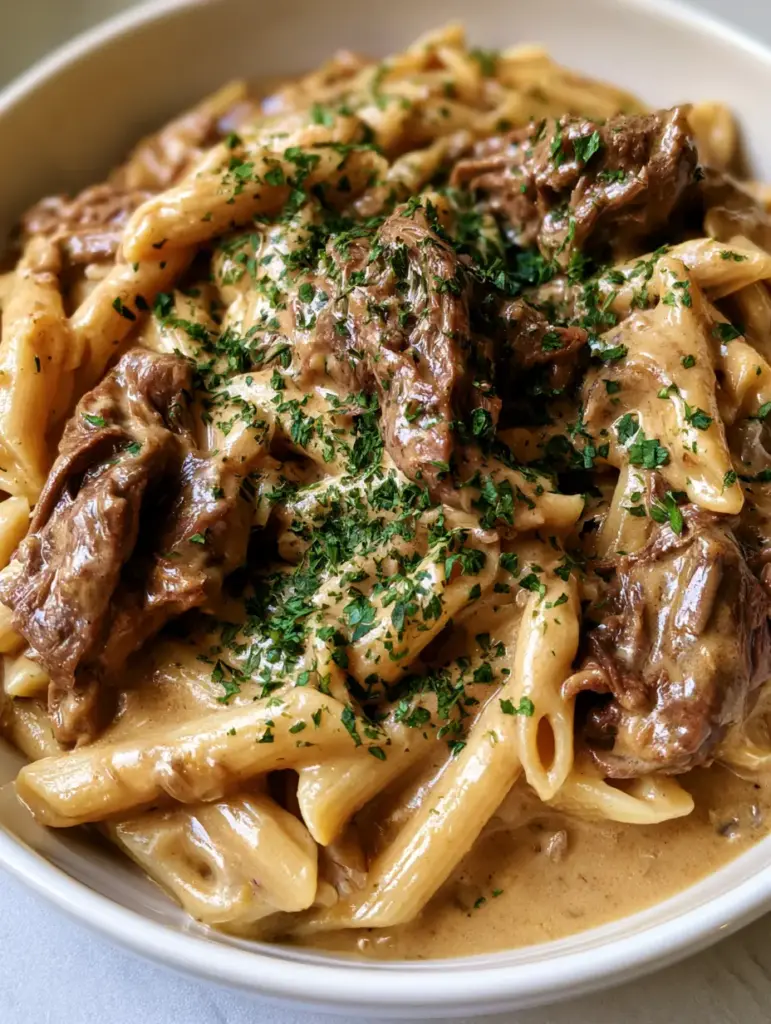 A white ceramic bowl filled with al dente penne and shredded beef chunks coated in garlic cream sauce.