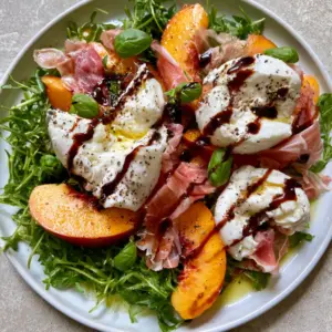 Top-down view of Peach Burrata Salad on a white ceramic plate with fresh arugula and a mahogany balsamic drizzle.