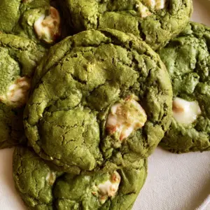 Several thick soft-baked matcha cookies stacked naturally on a neutral surface, showing deep green hue and caramelized white chocolate edges. (Matcha White Chocolate Cookies)